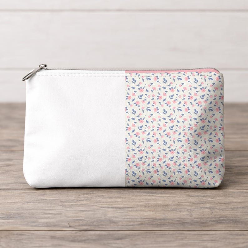 May include: A white zippered pouch with a floral pattern on the right side. The pattern features small pink flowers and blue leaves on a cream background. The pouch has a silver zipper.