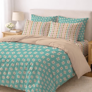 Op de afbeelding: Een bed met een bloemenpatroon dekbedovertrek en kussenslopen. Het dekbedovertrek is turquoise met witte bloemen, terwijl de kussenslopen een rechthoekig patroon in verschillende kleuren hebben. Het bed heeft ook een beige hoeslaken.