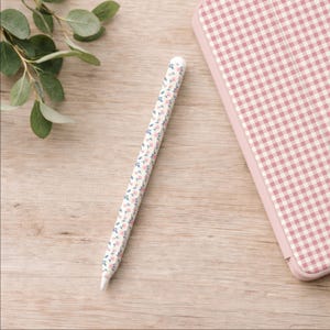 May include: A white stylus with a floral pattern of pink and blue flowers and green leaves. The stylus is next to a pink and white checkered tablet case on a light brown wooden surface. Green leaves are in the background.