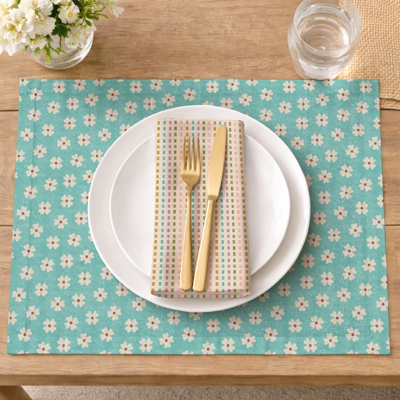Op de afbeelding: Een placemat met een turquoise achtergrond en een patroon van kleine witte bloemen met rode centra. Een wit bord, gouden vork en mes en een patroon servet liggen erop. Een glas water en een vaas met witte bloemen zijn ook aanwezig.