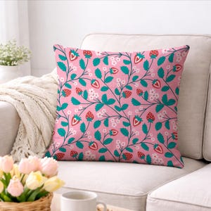 May include: Square throw pillow with a pink background and a repeating pattern of strawberries, green leaves, and white flowers. The pillow is on a white couch. The strawberries are red with green tops.