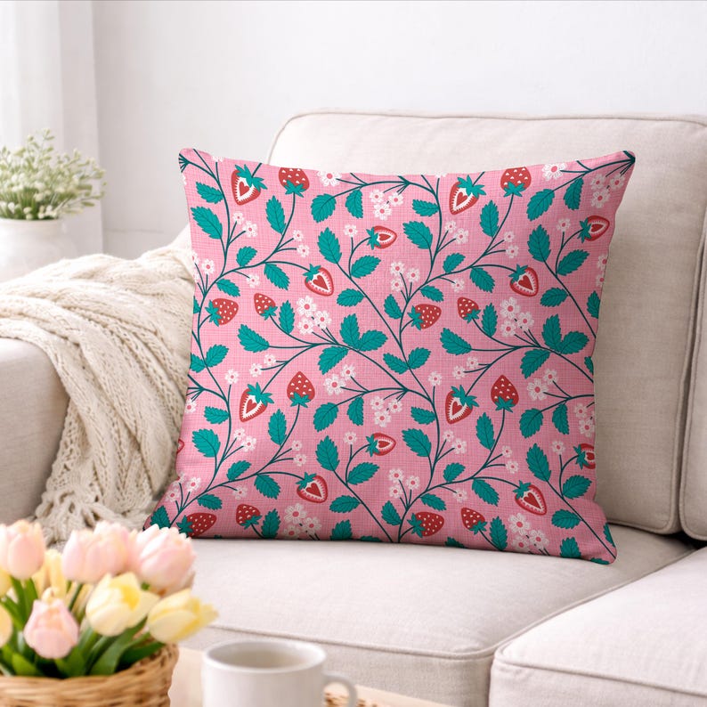 May include: Square throw pillow with a pink background and a repeating pattern of strawberries, green leaves, and white flowers. The pillow is on a white couch. The strawberries are red with green tops.