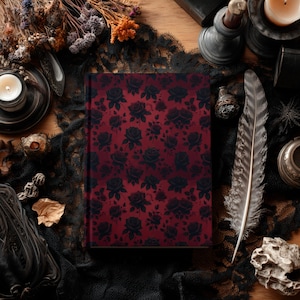 May include: A hardcover journal with a dark red cover featuring a pattern of black roses. The journal is surrounded by gothic-themed decor, including a feather, candles, and dried flowers, creating a vintage aesthetic.