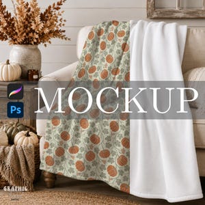 May include: A soft throw blanket featuring a repeating pattern of orange pumpkins and sage green leaves on a light cream background. The blanket is draped over a sofa, with one side showing the patterned fabric and the other side a solid white.