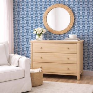 Op de afbeelding: Een licht houten dressoir met drie lades, een ronde spiegel met een geweven frame en een witte fauteuil. Het blauw-witte behang met patroon voegt een decoratief tintje toe. Een vaas met witte bloemen staat op het dressoir.