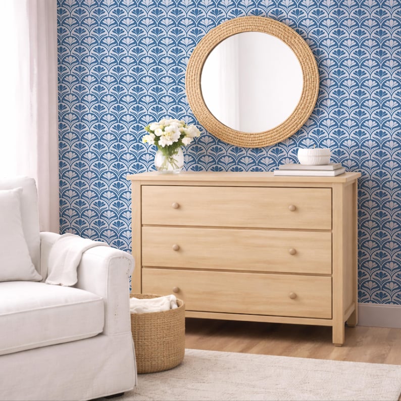 Op de afbeelding: Een licht houten dressoir met drie lades, een ronde spiegel met een geweven frame en een witte fauteuil. Het blauw-witte behang met patroon voegt een decoratief tintje toe. Een vaas met witte bloemen staat op het dressoir.