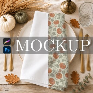 May include: A folded white cloth napkin featuring a vertical panel with a repeating pattern of orange pumpkins and green leaves on a light background. The napkin is placed on a round plate with gold cutlery, dried foliage, and small pumpkins.