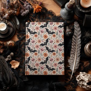 May include: A hardcover journal with a cream-colored background, featuring a repeating pattern of pink skulls, black bats, orange flowers, and small bones. The journal is closed and lies on a black lace cloth.