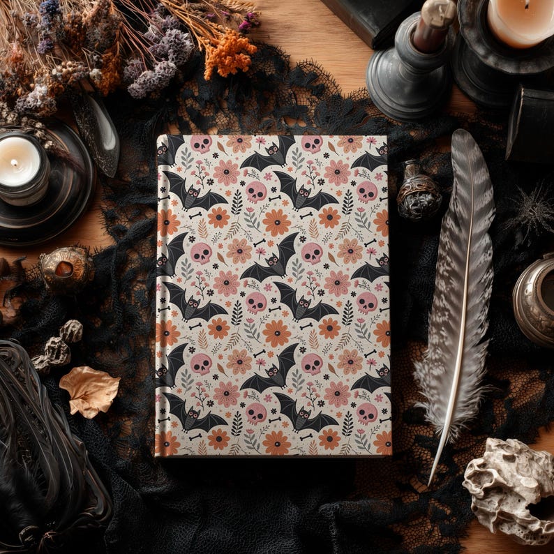 May include: A hardcover journal with a cream-colored background, featuring a repeating pattern of pink skulls, black bats, orange flowers, and small bones. The journal is closed and lies on a black lace cloth.