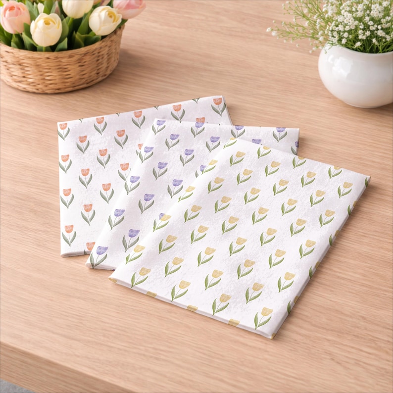 May include: Three white decorative napkins with a repeating pattern of watercolor tulips in orange, purple, and yellow. The napkins are folded and arranged on a light wooden surface, with a basket of tulips and a small vase of flowers in the background.