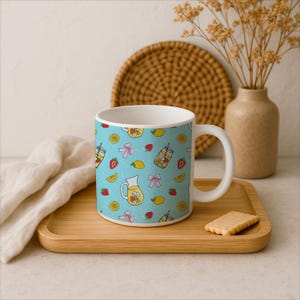 Puede incluir: Taza de cer&aacute;mica blanca con un patr&oacute;n azul claro de jarras de limonada, fresas, limones y lazos rosas. La taza est&aacute; sobre una bandeja de madera con una galleta.