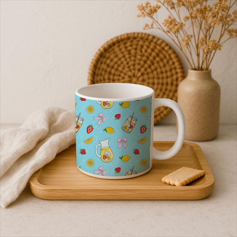 Puede incluir: Taza de cer&aacute;mica blanca con un patr&oacute;n azul claro de jarras de limonada, fresas, limones y lazos rosas. La taza est&aacute; sobre una bandeja de madera con una galleta.