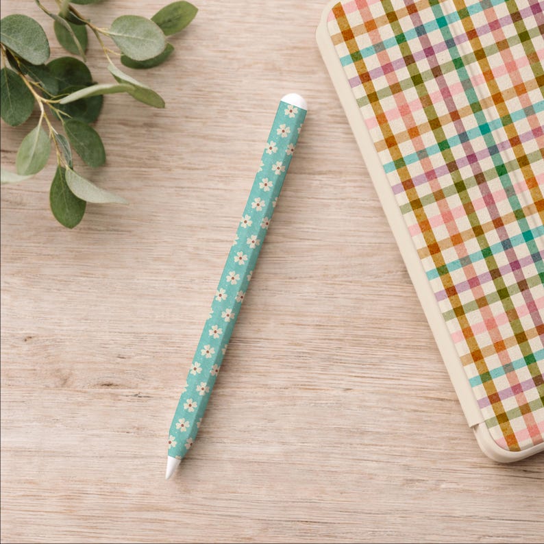 May include: A teal stylus pen with a white daisy pattern and a white tip. The pen is next to a notebook with a colorful plaid pattern. The background is a light wood surface with a sprig of green leaves.