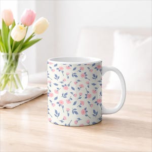 May include: White ceramic mug with a floral pattern of small pink flowers and blue leaves on a cream background. The mug has a white handle and is sitting on a wooden surface.