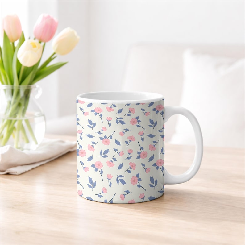 May include: White ceramic mug with a floral pattern of small pink flowers and blue leaves on a cream background. The mug has a white handle and is sitting on a wooden surface.
