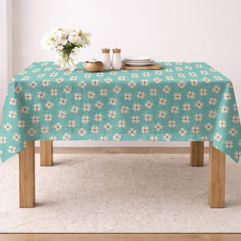 Op de afbeelding: Een rechthoekig turquoise tafelkleed met een herhalend patroon van witte bloemen met bruine centra. Het tafelkleed ligt op een houten tafel. Een vaas met witte bloemen, zout- en pepervaatjes en borden staan op de tafel.