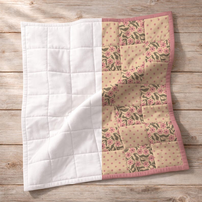 May include: A baby quilt with a patchwork design. One side is white, quilted in a grid pattern. The other side features a floral pattern with pink flowers and green leaves on a beige background. The quilt is trimmed with a pink border.