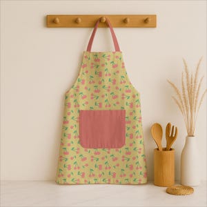 Puede incluir: Delantal amarillo con un estampado de cerezas rosas y un bolsillo rosa. El delantal cuelga de un perchero de madera. Tema de cocina y cocina.