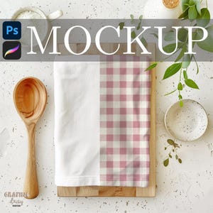 May include: A white and pink gingham tea towel rests on a wooden cutting board. A wooden spoon, small bowl, and greenery are arranged around the towel. The word "MOCKUP" is displayed across the top.