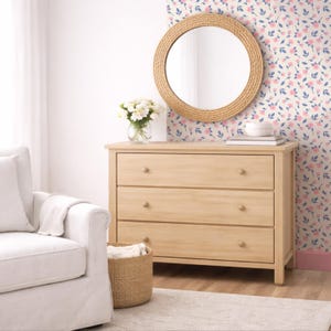 May include: A light wood three-drawer dresser with round wooden knobs. A round mirror with a woven frame hangs above the dresser. A vase of white flowers sits on top of the dresser. The wall behind the dresser has a floral pattern.