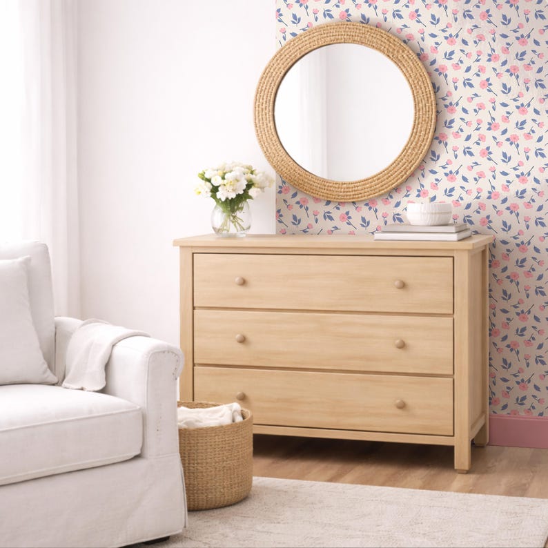 May include: A light wood three-drawer dresser with round wooden knobs. A round mirror with a woven frame hangs above the dresser. A vase of white flowers sits on top of the dresser. The wall behind the dresser has a floral pattern.