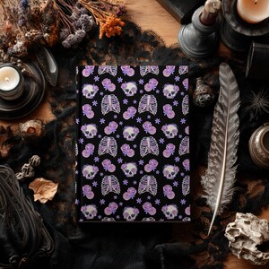 May include: A black journal with a repeating pattern of skulls, rib cages, purple roses, and stars. The journal is surrounded by gothic-themed items, including a feather, candles, and dried flowers.