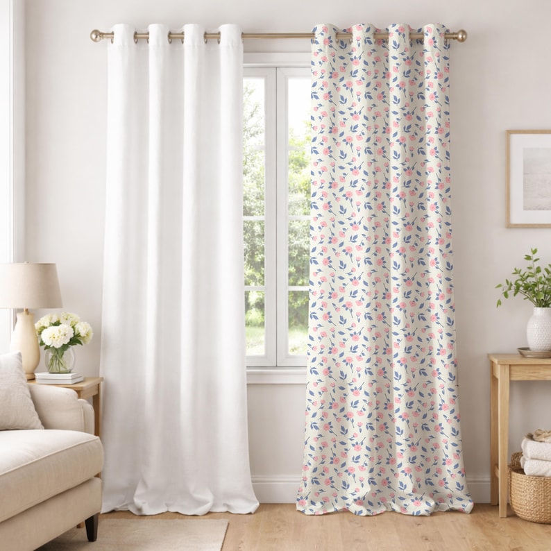 May include: Two window curtains. One is solid white, the other has a cream background with a floral pattern of pink flowers and blue leaves. The curtains are hanging on a gold rod with grommets. The room has a neutral color scheme.
