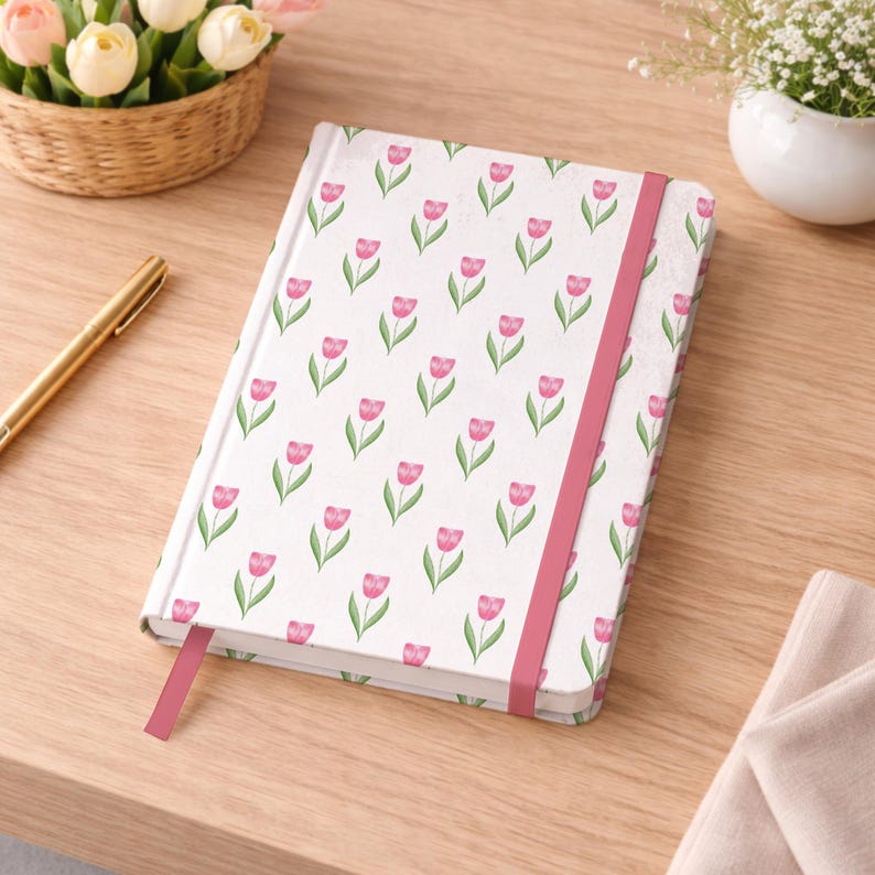 May include: A white notebook with a pink tulip pattern and a matching elastic band closure. The notebook is open on a wooden surface, with a gold pen and a basket of flowers in the background.