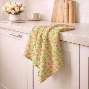May include: A yellow kitchen towel with a repeating pattern of pink cherries and green stems. The towel is draped over a white cabinet, with a countertop and a vase of flowers in the background. The towel is made of a soft, textured fabric.