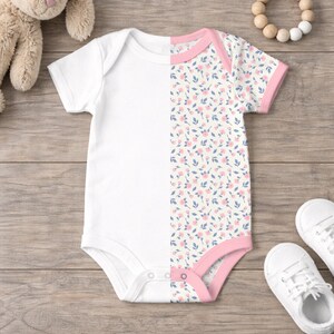 May include: A white baby onesie with a floral pattern on the right side. The floral pattern features small pink flowers and blue leaves on a cream background. The onesie has pink trim and snaps at the bottom.
