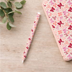 May include: A white stylus with a pink bow pattern and a matching tablet case. The bows are in shades of pink and red on a cream background. The stylus is on a light wood surface.