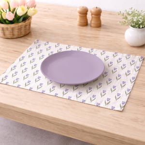 May include: A lavender plate rests on a cream-colored placemat adorned with a repeating pattern of purple tulip illustrations. The placemat is on a light wooden table, with a basket of flowers and other decorative items in the background.