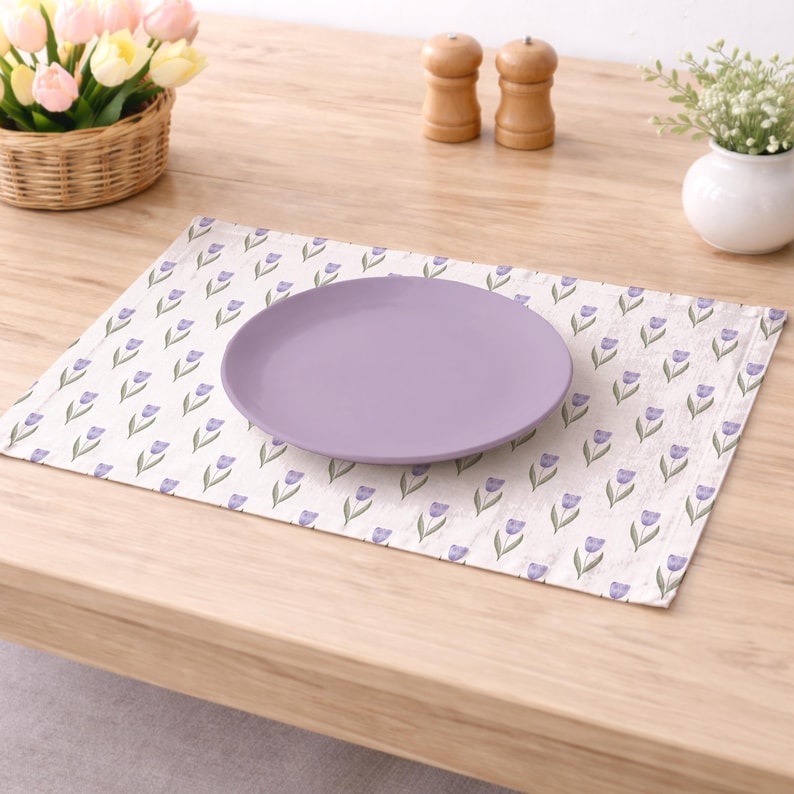 May include: A lavender plate rests on a cream-colored placemat adorned with a repeating pattern of purple tulip illustrations. The placemat is on a light wooden table, with a basket of flowers and other decorative items in the background.