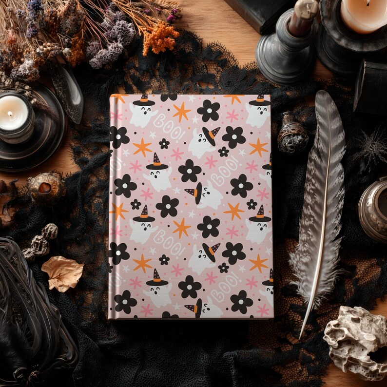 May include: A pink journal with a Halloween-themed pattern featuring white ghosts wearing witch hats, black flowers, orange stars, and the word "BOO!". The journal is surrounded by decorative items.