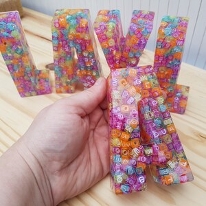 May include: Colorful resin letters spelling "LAVA" with a rainbow of small plastic beads embedded in the resin. The letters are translucent and the beads are visible through the resin.