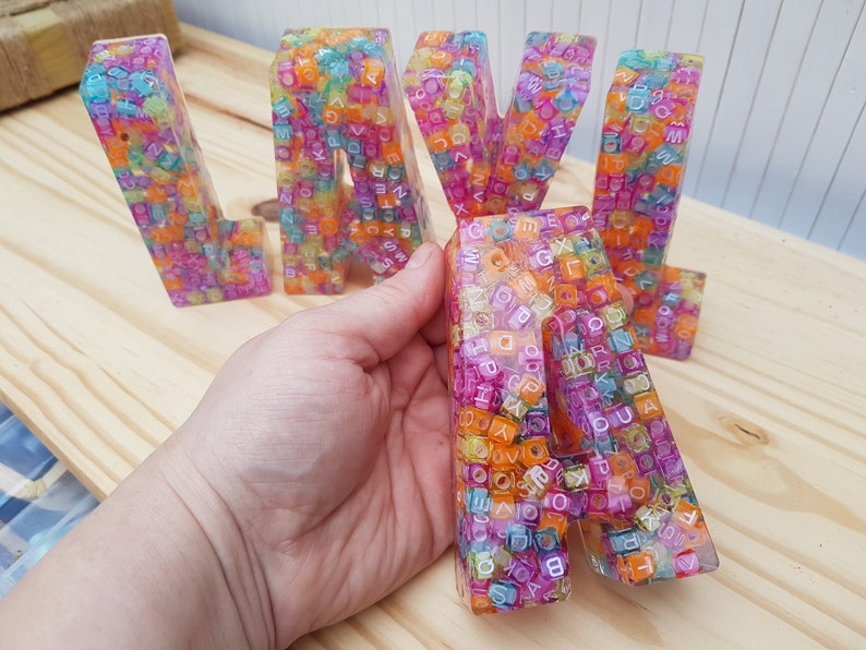 May include: Colorful resin letters spelling "LAVA" with a rainbow of small plastic beads embedded in the resin. The letters are translucent and the beads are visible through the resin.