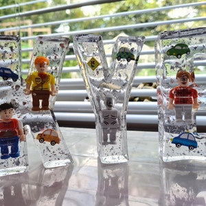 May include: Four clear resin letters spelling out the name "KYLE" with small toy figures and cars embedded inside. The letters are sitting on a white surface.