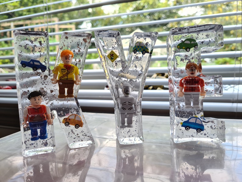 May include: Four clear resin letters spelling out the name "KYLE" with small toy figures and cars embedded inside. The letters are sitting on a white surface.