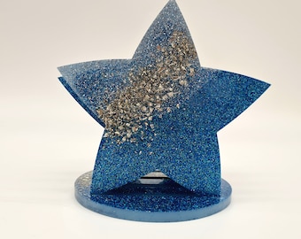 Puede incluir: Un porta servilletas en forma de estrella azul con purpurina y detalles de purpurina plateada. El porta servilletas tiene una base redonda.