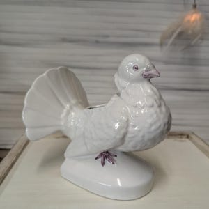 Può includere: Figurina di colomba in ceramica bianca con un design dipinto di viola sui piedi. La colomba ha una coda rialzata ed è posata su una base ovale bianca.