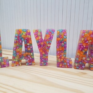 May include: Colorful, multi-colored, resin letters spelling out the name "LAYLA". Each letter is made of small, colorful, plastic beads with letters on them.