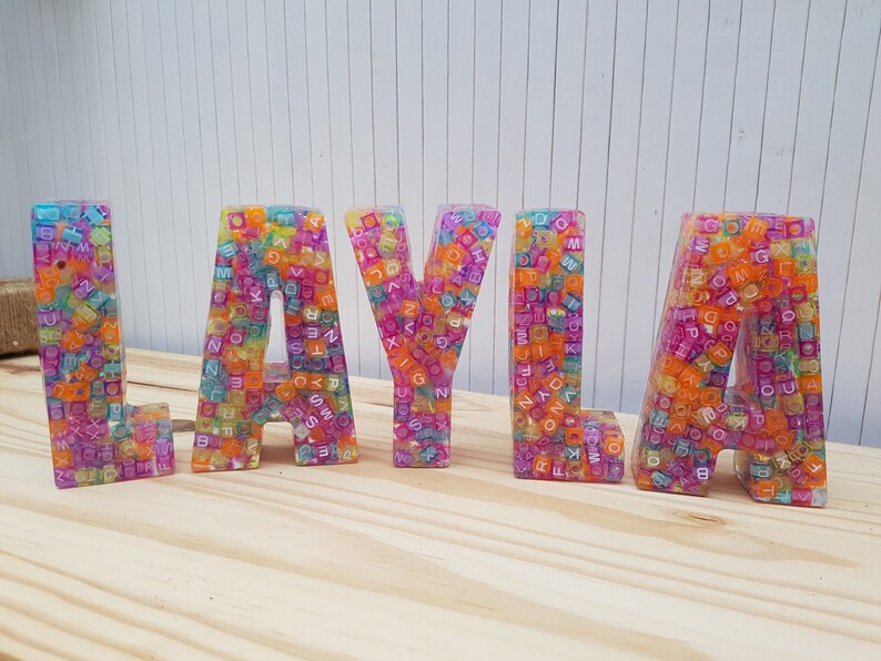 May include: Colorful, multi-colored, resin letters spelling out the name "LAYLA". Each letter is made of small, colorful, plastic beads with letters on them.