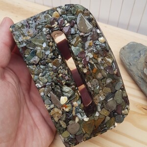 May include: A letter D made of resin with embedded small stones in shades of brown, green, and gray. The letter has a slot cut out of the center.