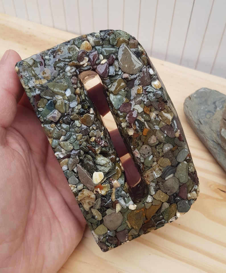May include: A letter D made of resin with embedded small stones in shades of brown, green, and gray. The letter has a slot cut out of the center.