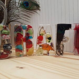 May include: The word "CRAFT" spelled out in colorful resin letters. Each letter is filled with different materials, including feathers, flowers, and fabric.