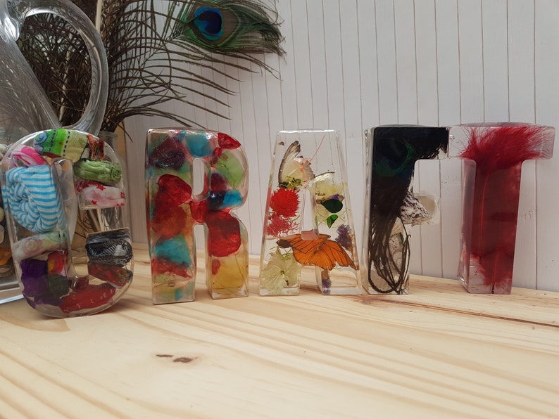 May include: The word "CRAFT" spelled out in colorful resin letters. Each letter is filled with different materials, including feathers, flowers, and fabric.