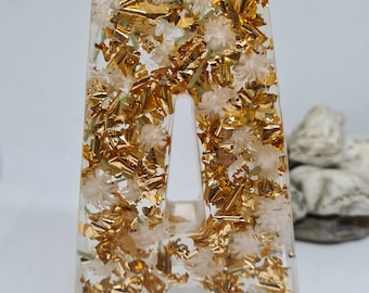 Könnte beinhalten: Ein Harzbuchstabe A mit goldenen Flocken und weißen Blumen, die darin eingebettet sind. Der Buchstabe ist klar und die goldenen Flocken und Blumen sind im gesamten Buchstaben sichtbar.