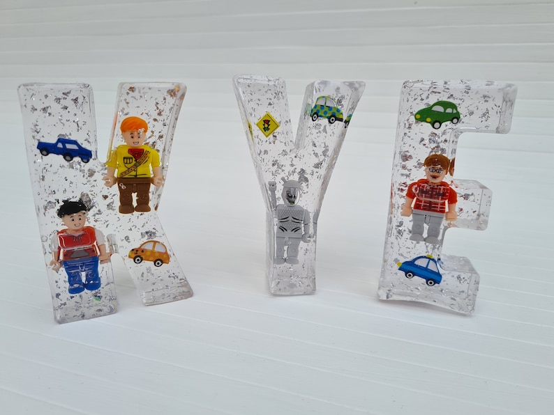 May include: Clear resin letters spelling the name "KYE" with small toy figures and cars embedded inside. The letters are filled with silver glitter.