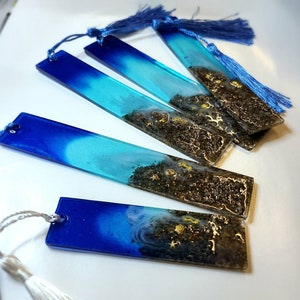 Könnte beinhalten: Vier blaue und türkisfarbene Lesezeichen aus Harz mit goldenem Glitzer und einer schwarzen sandartigen Textur. Jedes Lesezeichen hat einen Quasten.