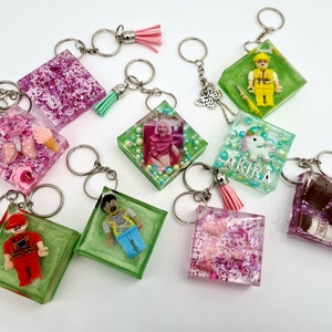 Peut inclure: Neuf breloques de porte-clés en résine aux thèmes variés. Les breloques sont carrées et présentent des paillettes, de petites figurines et d'autres éléments décoratifs. Certaines breloques ont un pompon attaché au porte-clés.